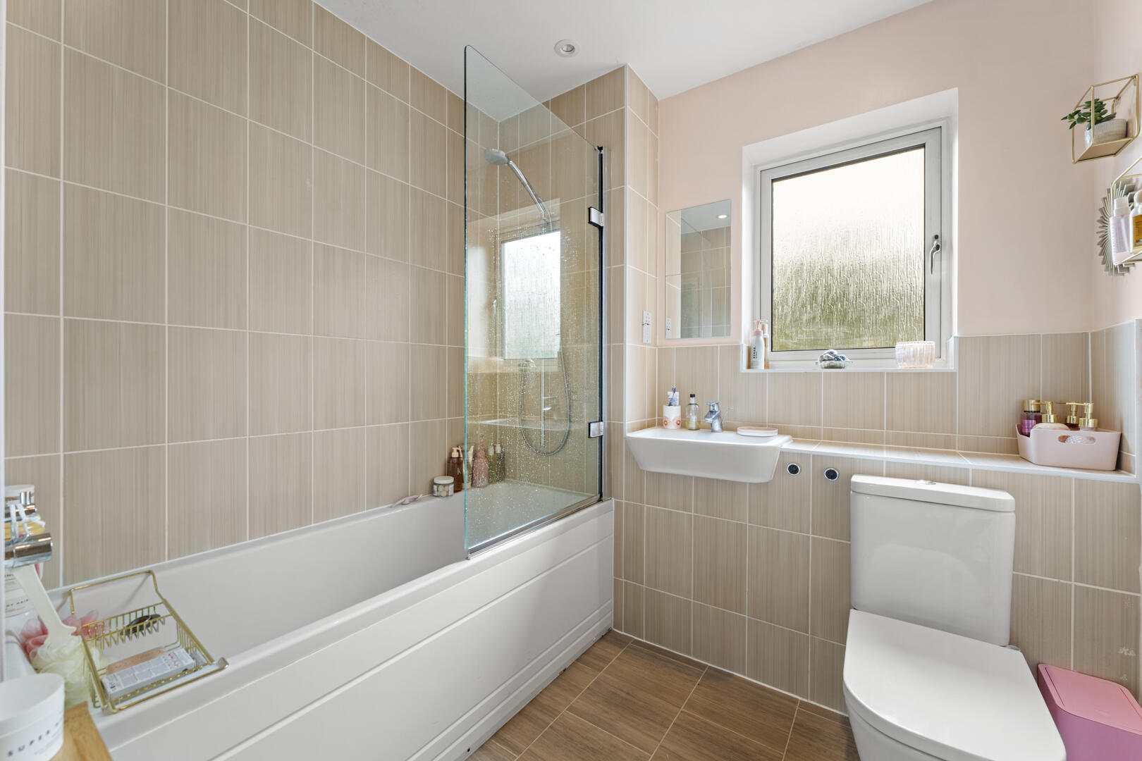 Bright beige tiled bathroom with a white bathtub, glass shower screen, sink, and a toilet beneath a frosted window.
