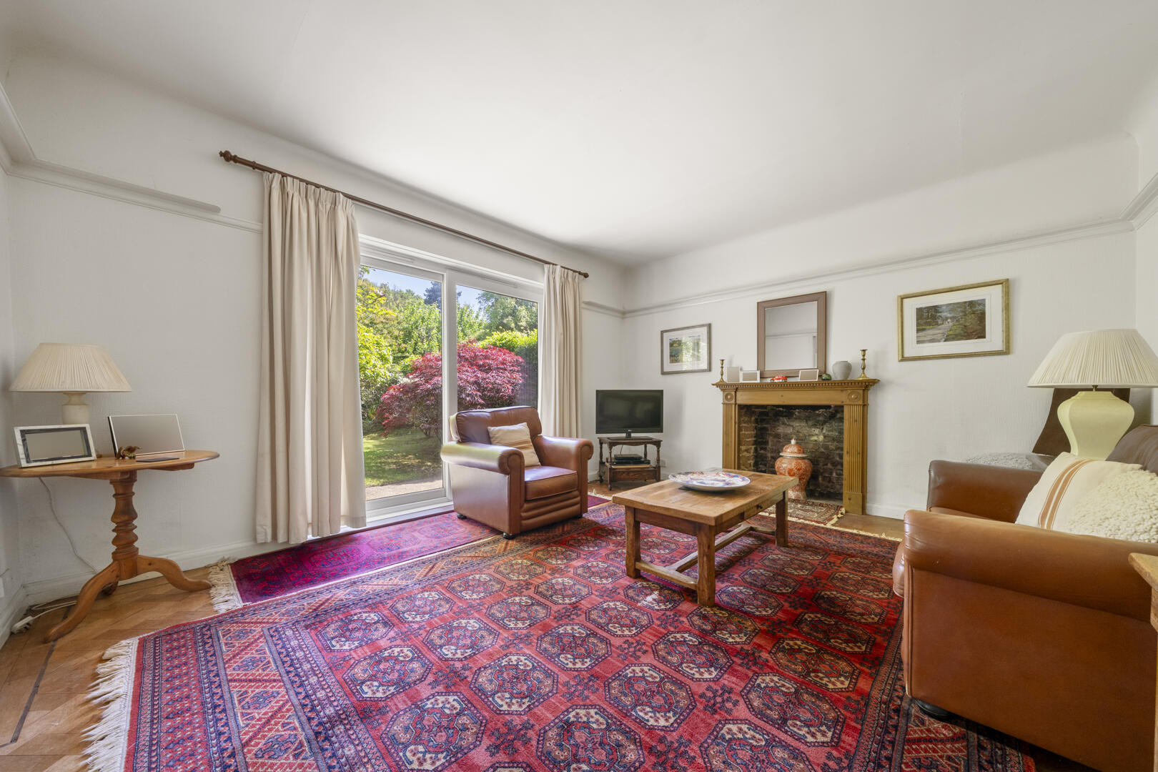 Living room with a red patterned rug, brown leather chair, coffee table, TV, and a stone fireplace; sliding glass door to a green garden outside.