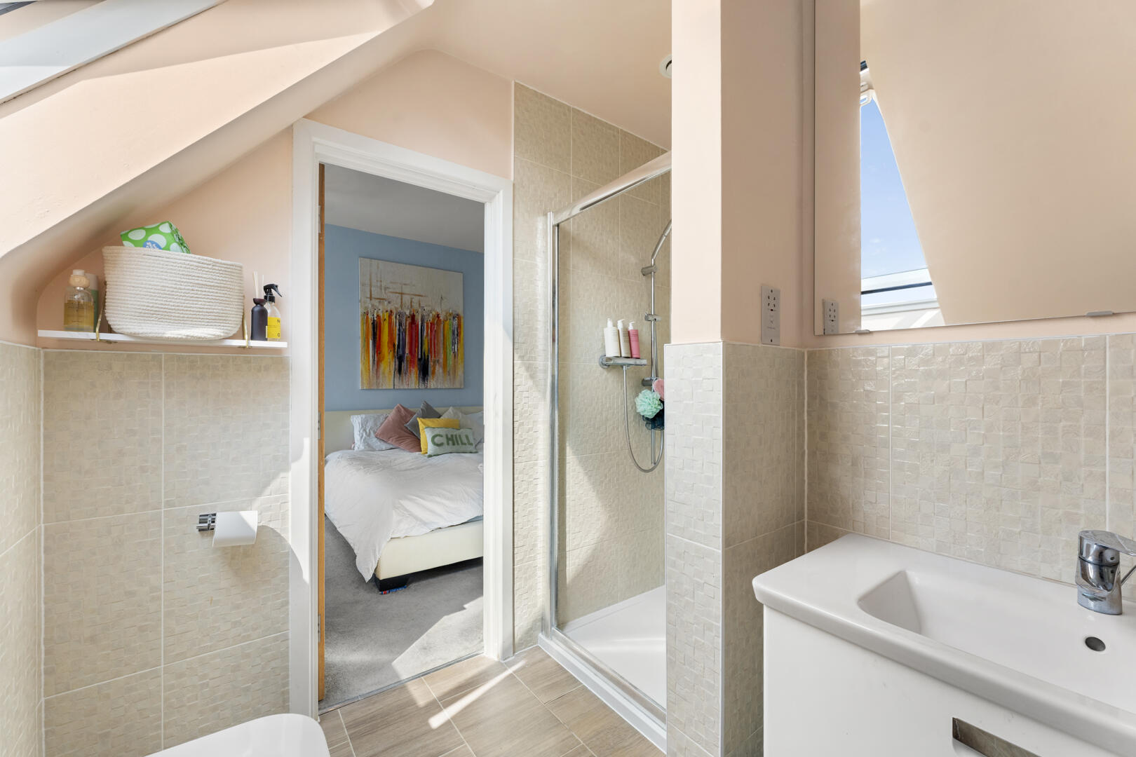 Small bathroom with a glass shower, sink, and toilet, open to a sunny bedroom with a colorful wall art piece seen through the doorway—slanted ceiling above.