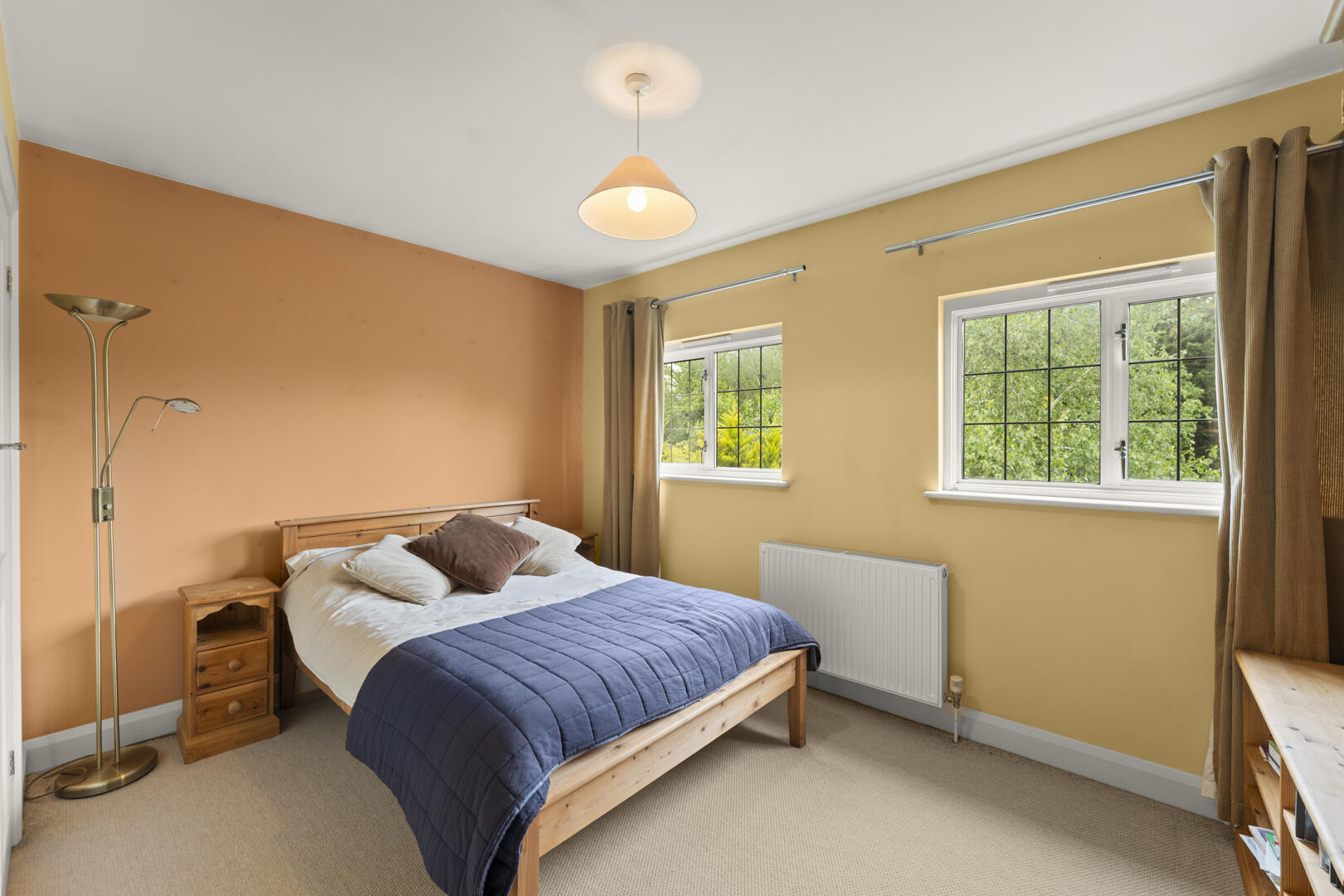 Cozy bedroom with a wooden bed, navy comforter, and beige carpet; two windows with beige curtains on mustard walls