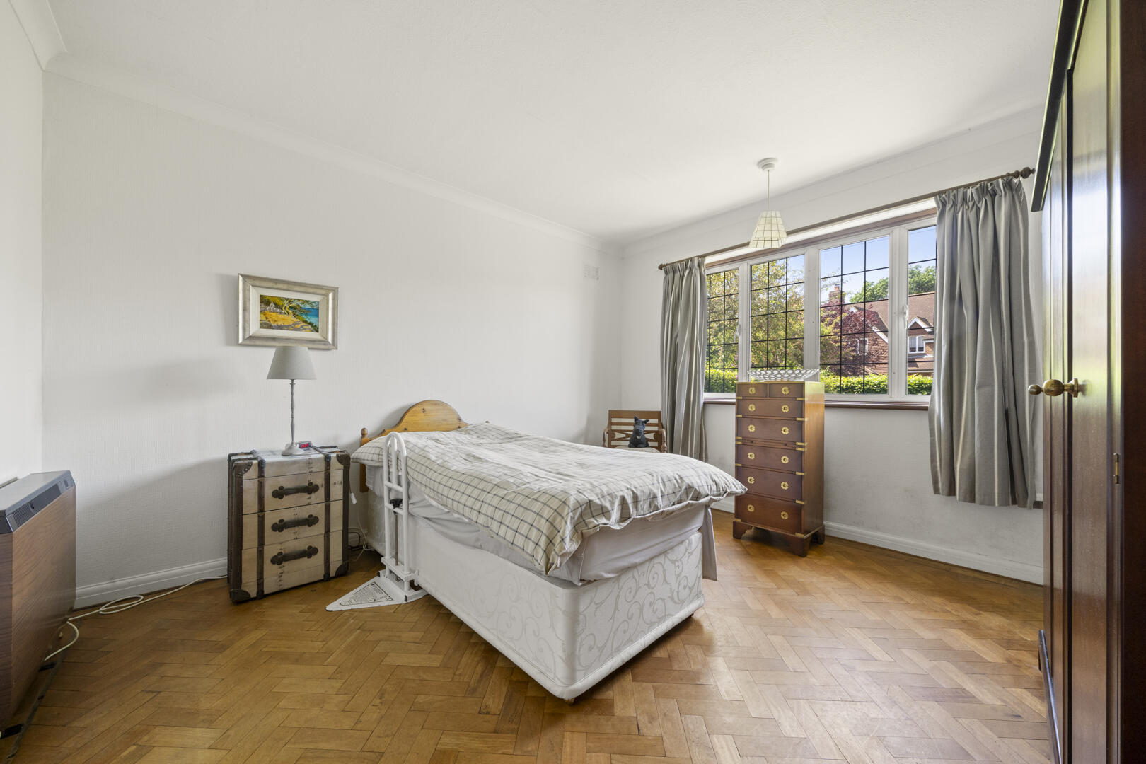 A tidy bedroom with a single bed, plaid duvet, vintage trunk bedside table, and a tall chest of drawers by a window with gray curtains.