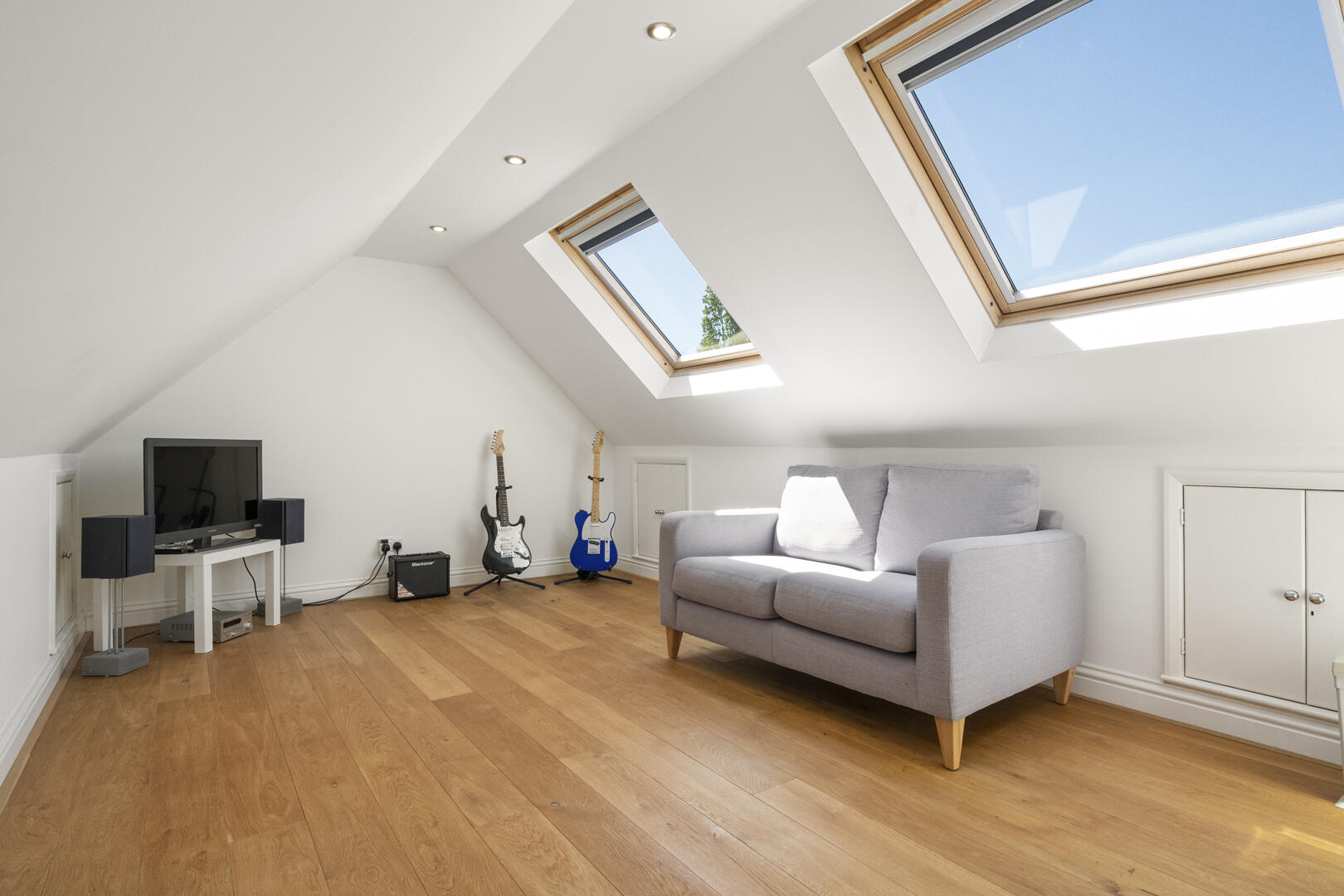 Bright attic living room with two skylights, a gray sofa, and a wooden floor; guitars on stands, TV and speakers in a small media setup nearby.