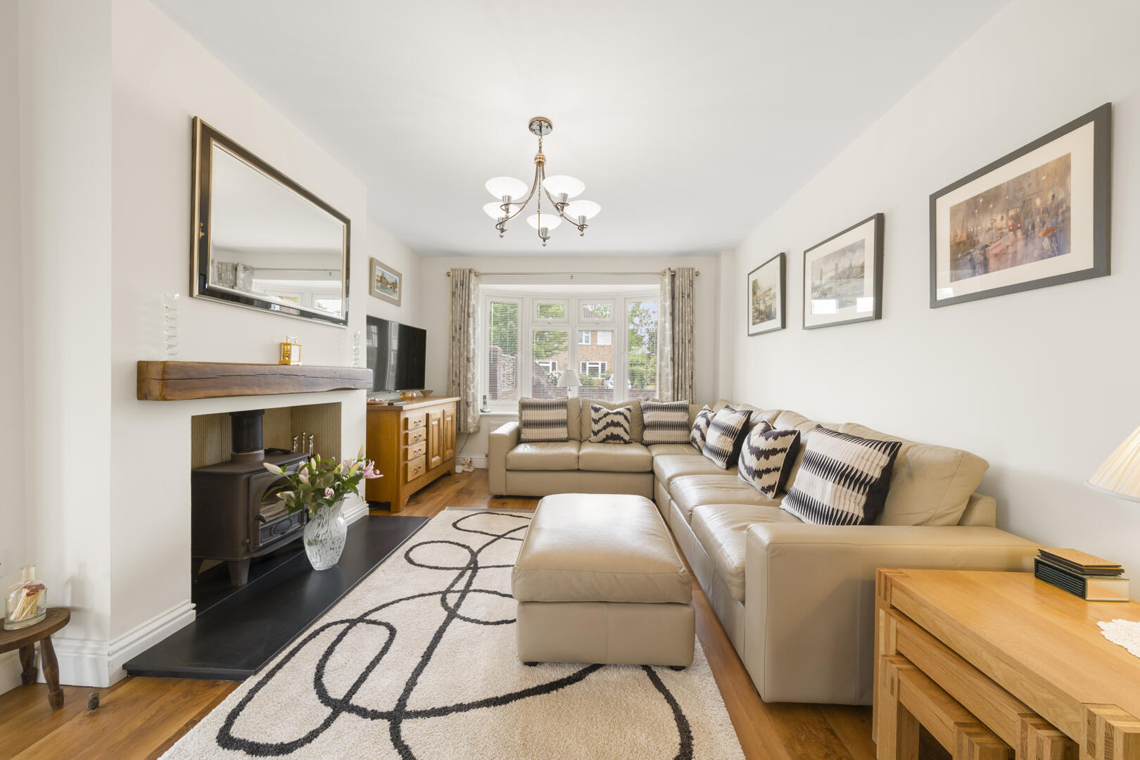 Bright living room with a beige L-shaped sofa, matching ottoman, and patterned cushions facing a windowed bay; chandelier overhead and framed art along the wall.