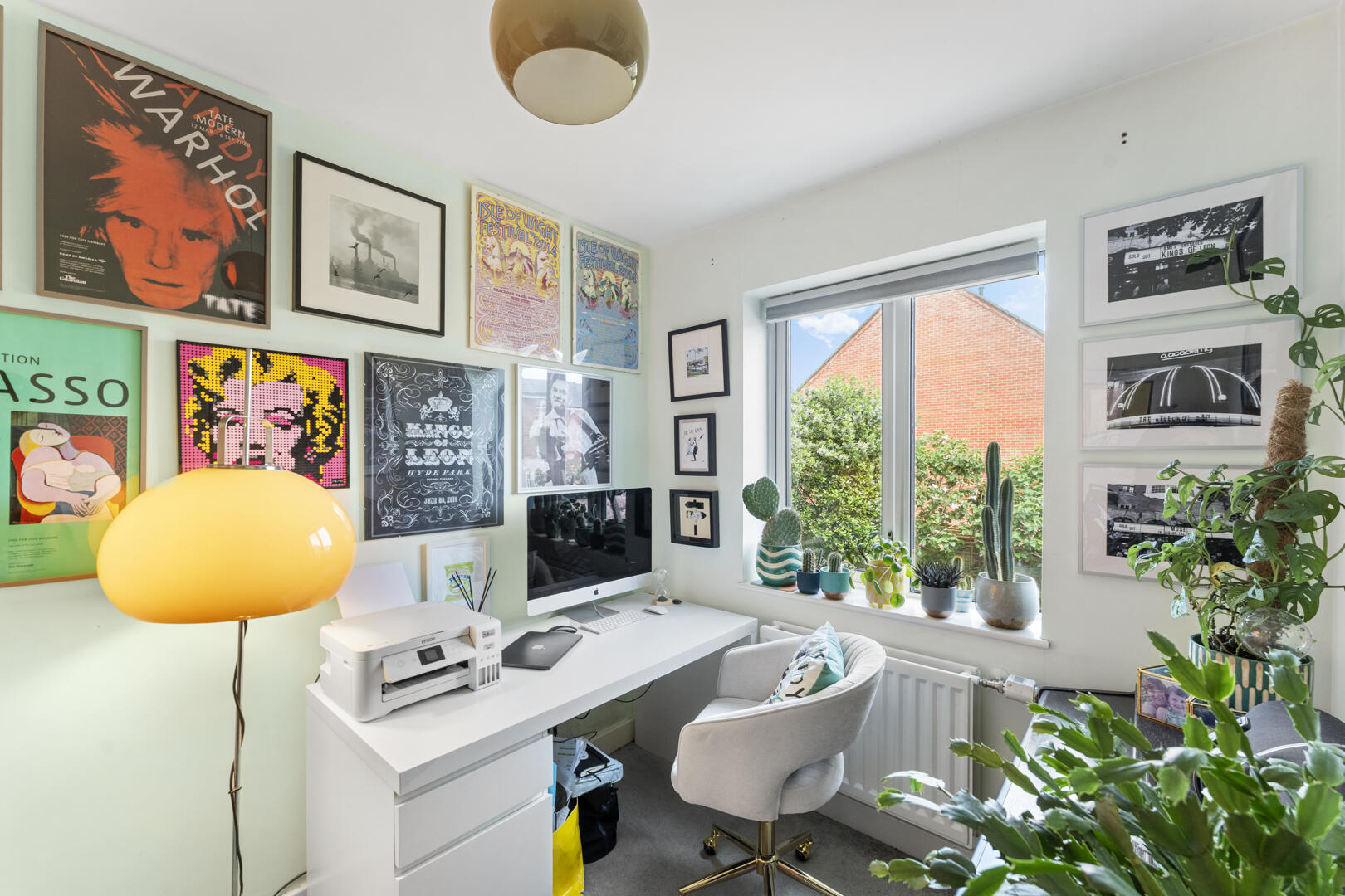 Bright home office with a wall of colorful posters, a white desk holding an iMac and printer, and potted plants by a large window.
