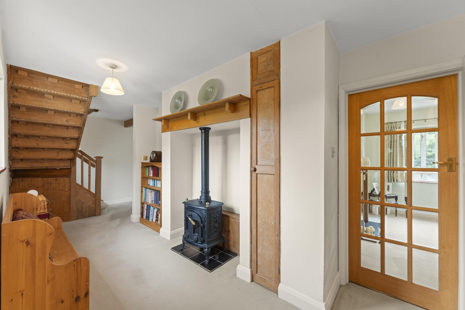 Living area with a black wood stove in a niche, wooden staircase on the left, and a wooden door to a sunlit room on the right