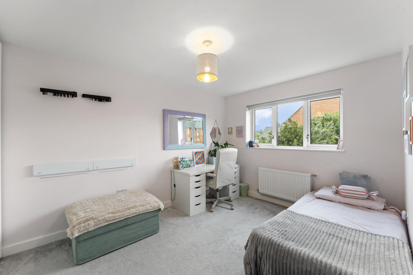 Bright, tidy bedroom with a white study desk, lavender-framed mirror, white chair, and a window looking out to trees and houses outside.