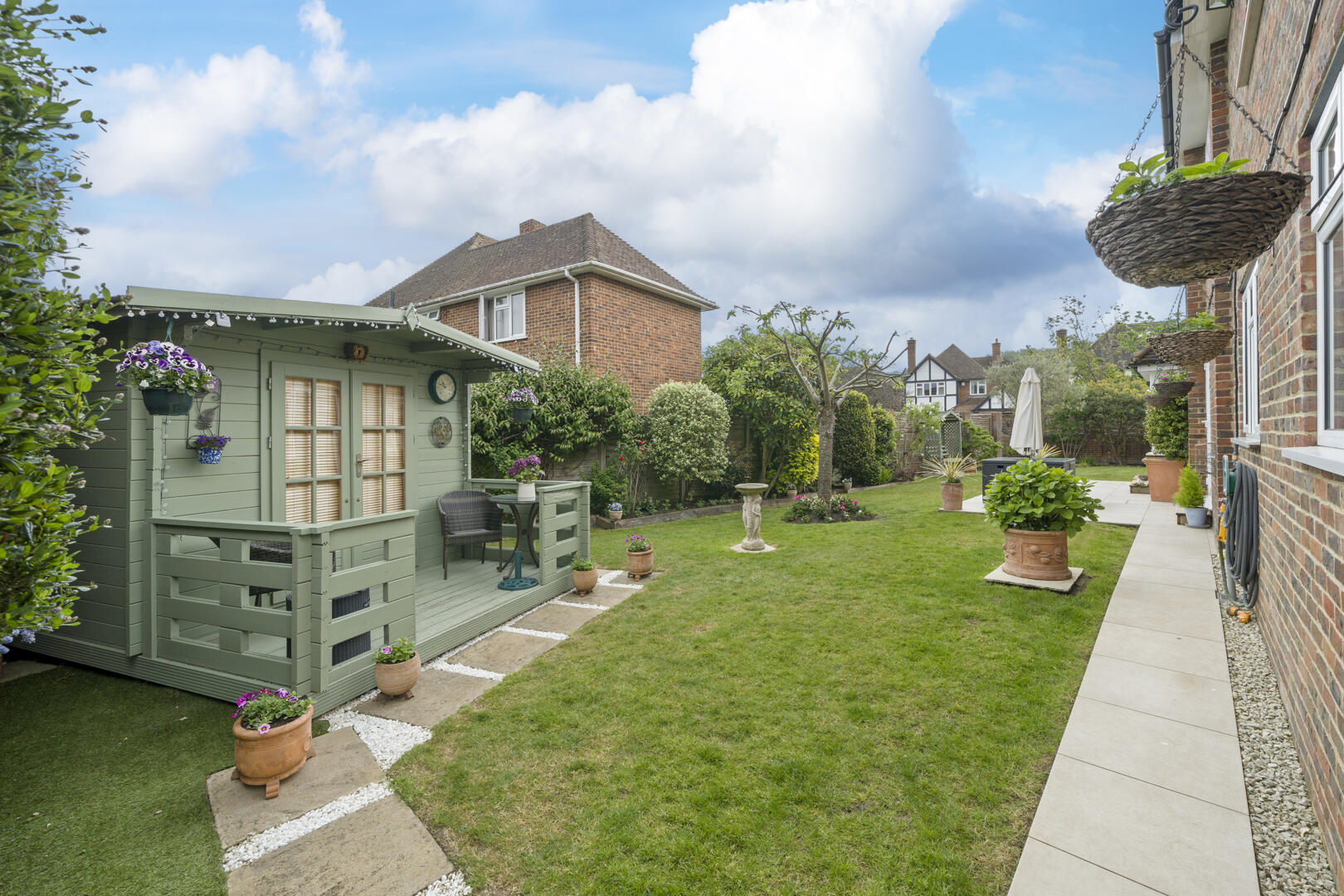Backyard with a green wooden shed on the left, small porch, potted flowers, and a well-manicured lawn beside a brick house.