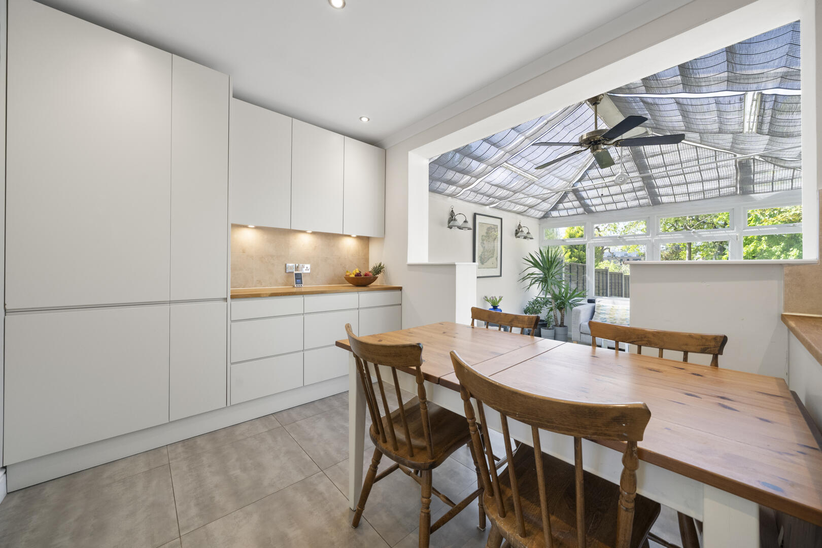 Modern white kitchen with tall cabinets, wooden dining table and chairs, opening to a sunlit conservatory with a ceiling fan.