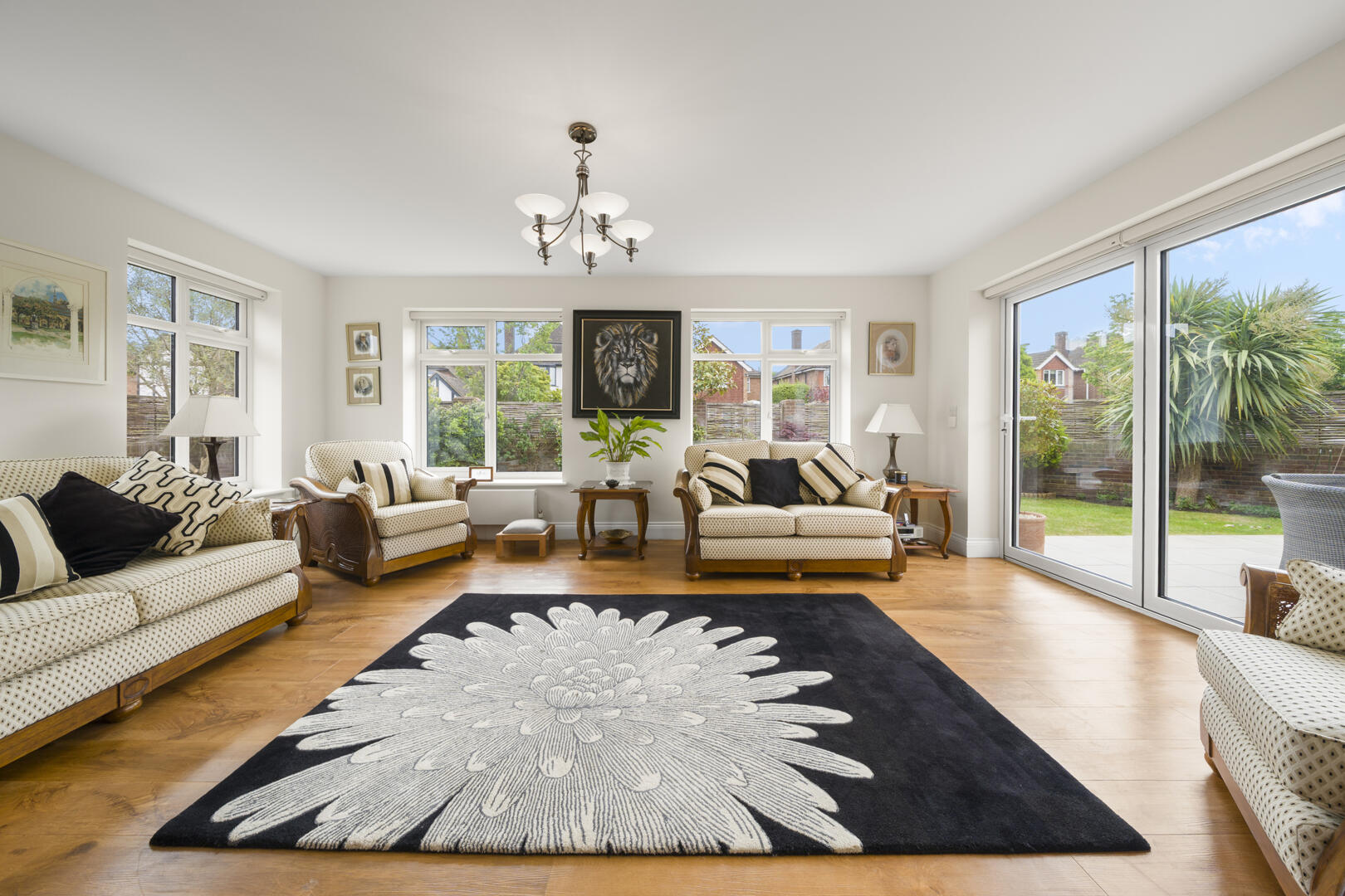 Bright living room with beige patterned sofas, a black rug with a large white flower, and warm wood flooring near big windows.