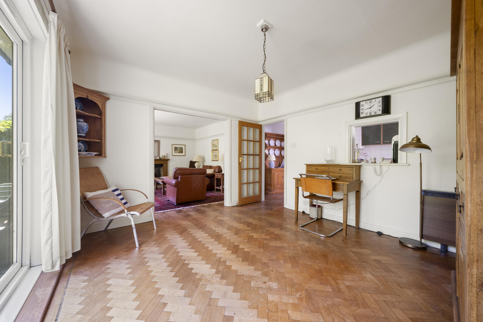 Spacious living room with chevron-patterned hardwood floor, a wooden desk and chair against the right wall, and doorways to other rooms ahead.