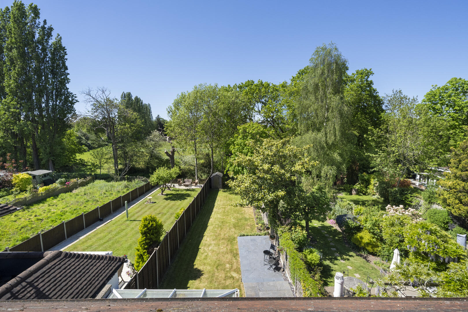 Aerial view of a sunny backyard with a manicured lawn, wooden fence, seating area, and tall trees.