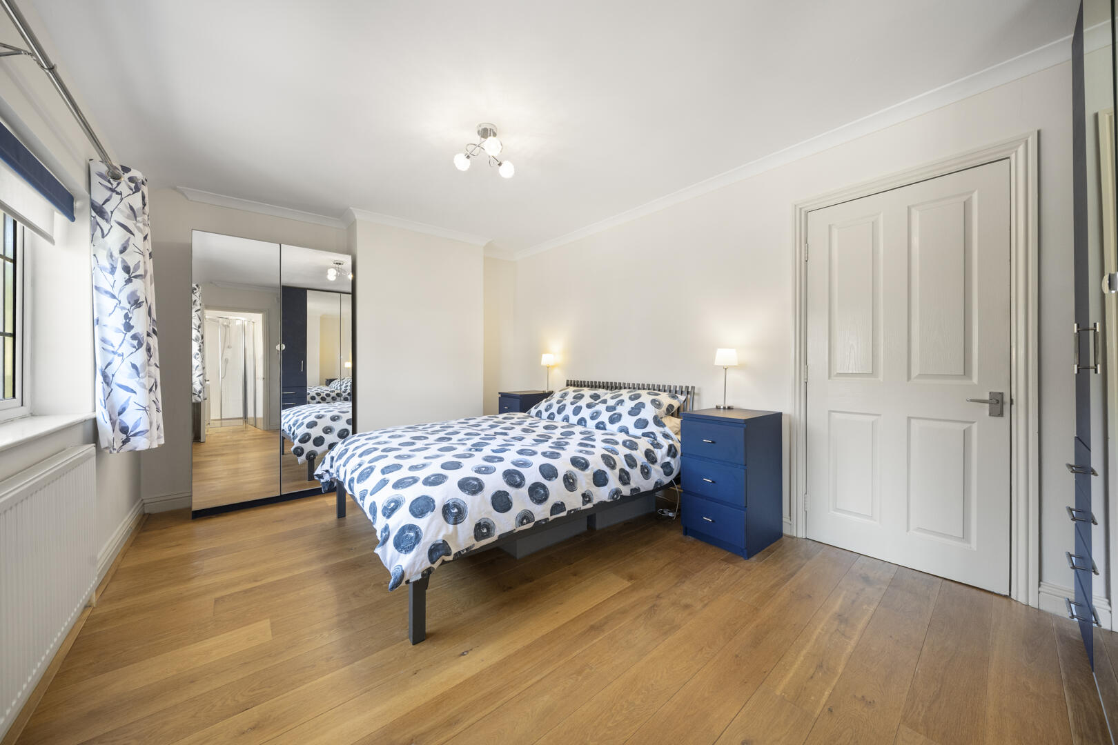 Bright bedroom with a double bed, blue nightstands, and a mirrored wardrobe; white walls and a polka-dot blue-and-white duvet.