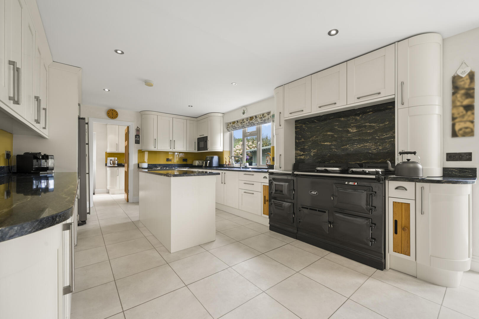Spacious white kitchen with a black Aga range, central island, white cabinets, and a yellow backsplash by the window.
