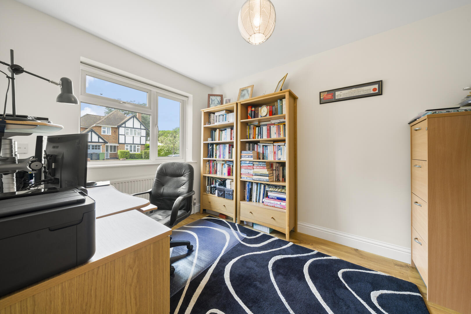 Home office with a desk, printer, black swivel chair, and two tall wooden bookshelves filled with books near a large window showing houses outside.