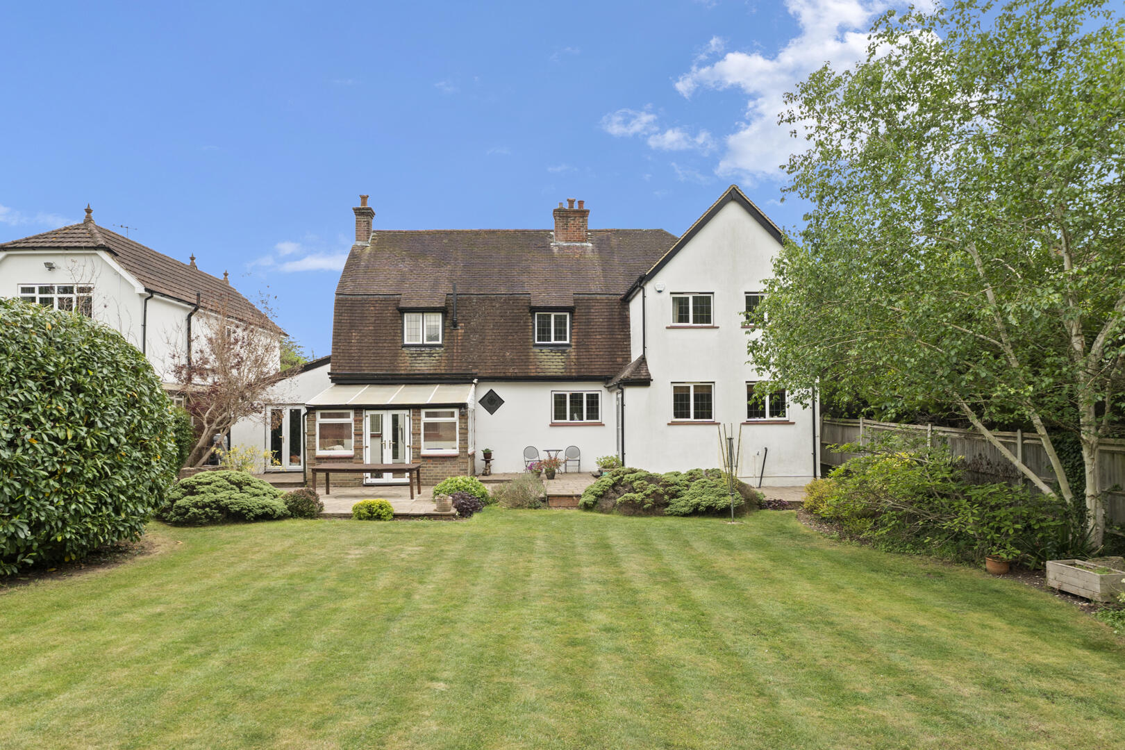 Backyard of a white two-story house with a brown-tiled roof, patio seating, and a well-kept lawn under a blue sky.