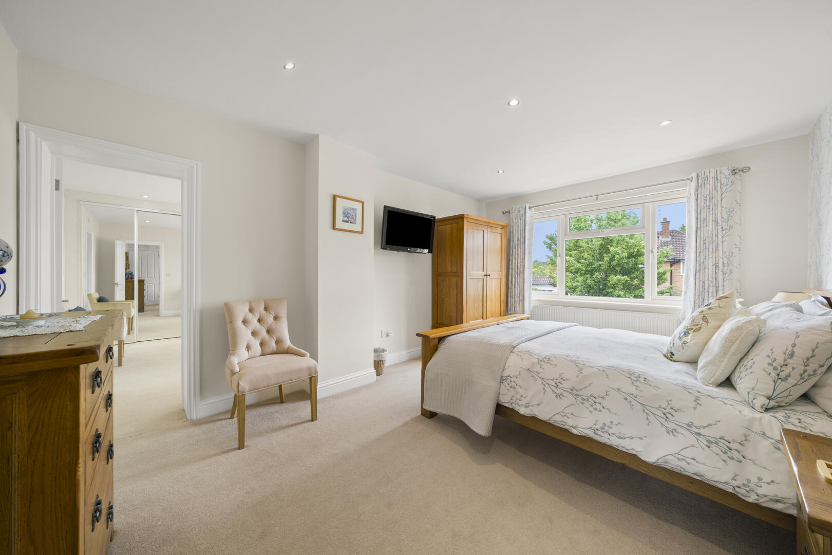 Bright master bedroom with a wooden bed, floral bedding, and a large window overlooking trees; wardrobe, wall TV, and beige carpeted floor nearby.