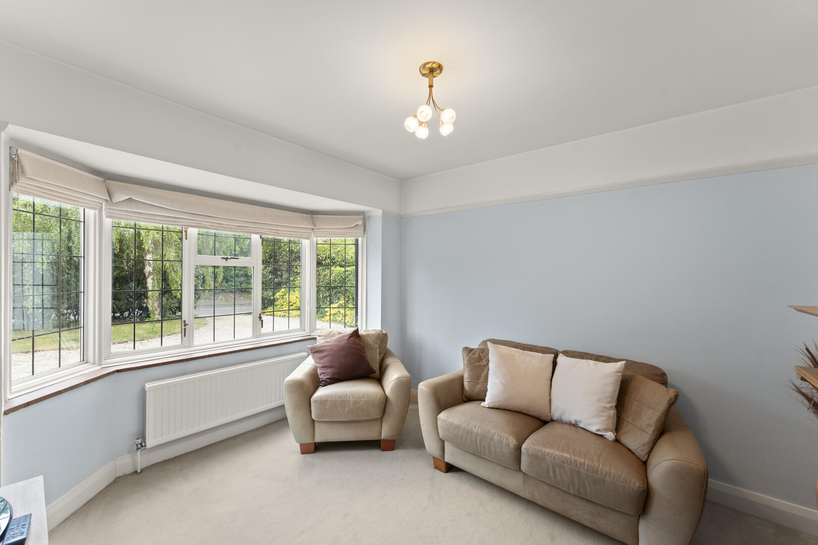 Living room with a beige sofa and armchair facing a bay window, light blue walls, and a small radiator beneath the window.