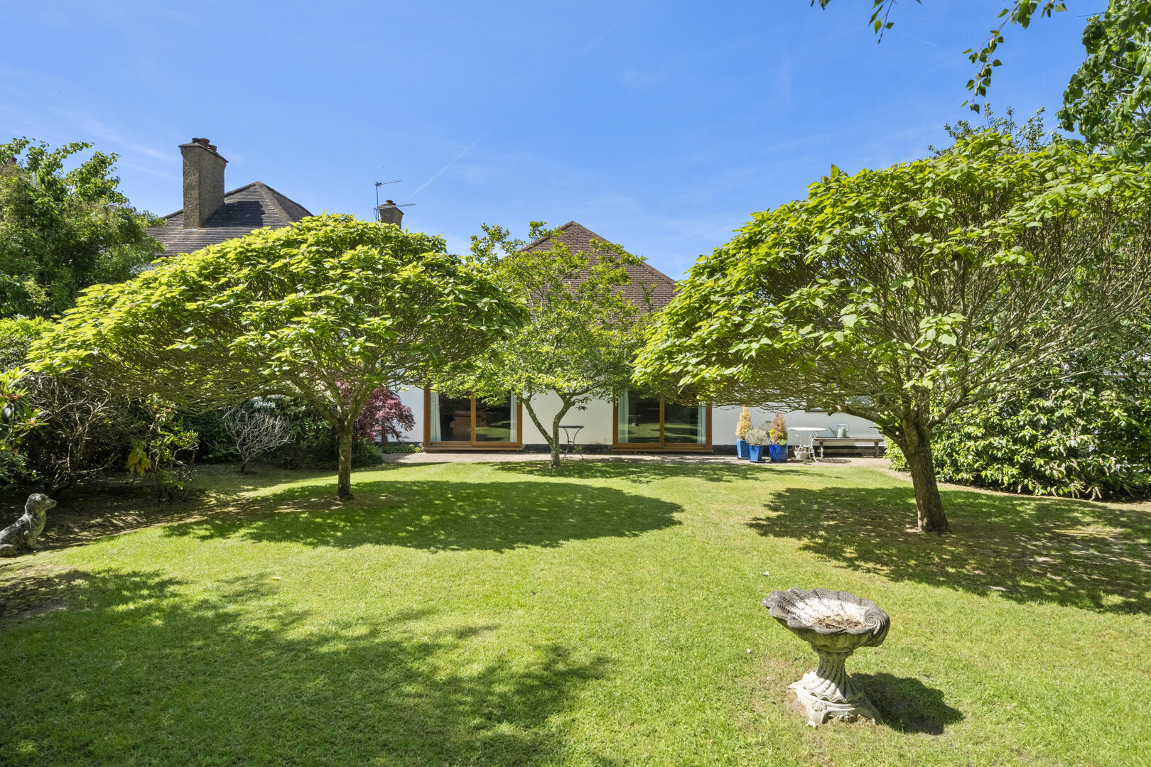 Sunny backyard with a well-kept lawn, two shade trees, and a single-story house with large glass doors in the background.