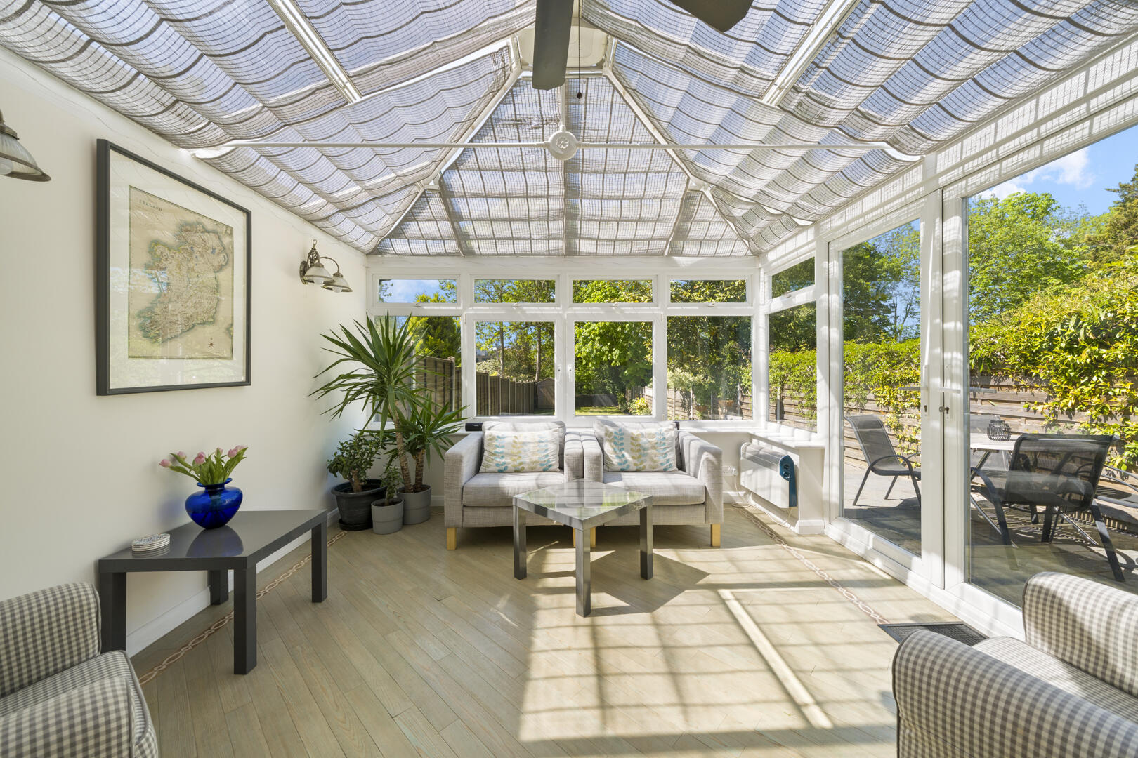 Bright conservatory living space with two gray sofas, a glass coffee table, and large windows revealing a leafy garden outside.