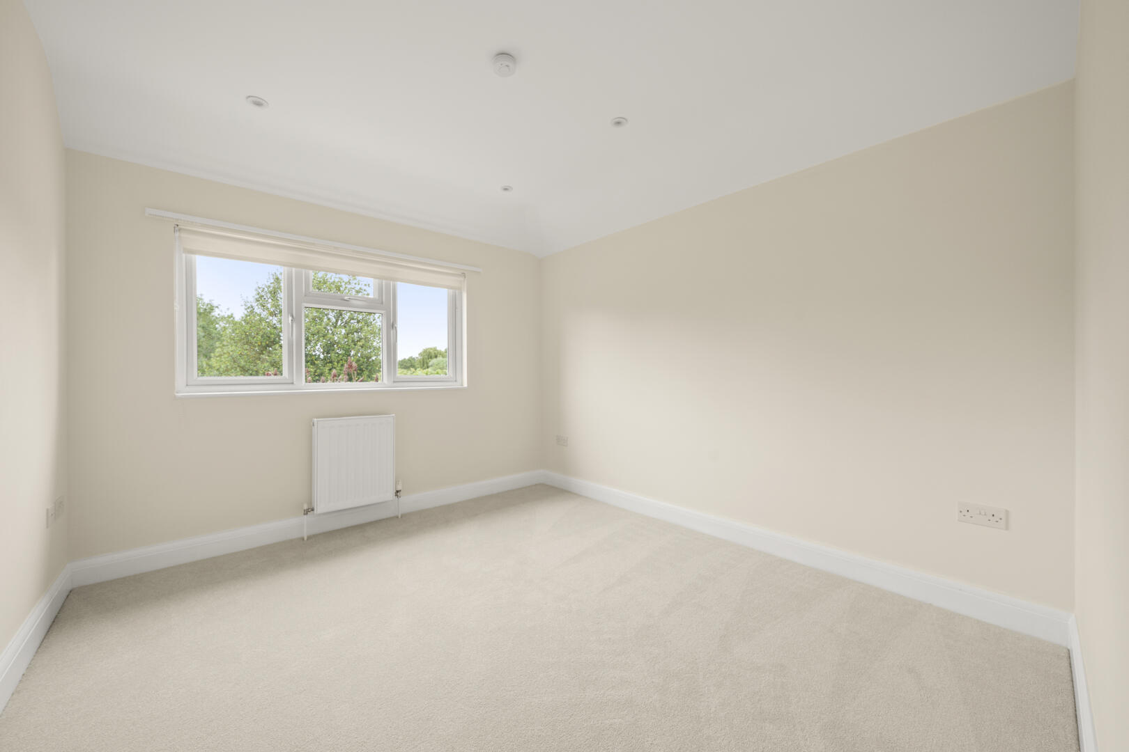 Empty beige bedroom with a large window, radiator beneath it, and cream carpet.
