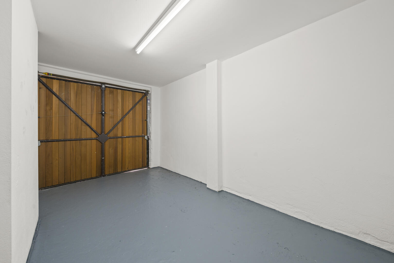 Empty garage with a wooden sectional door, white walls, and a gray concrete floor under a fluorescent ceiling light