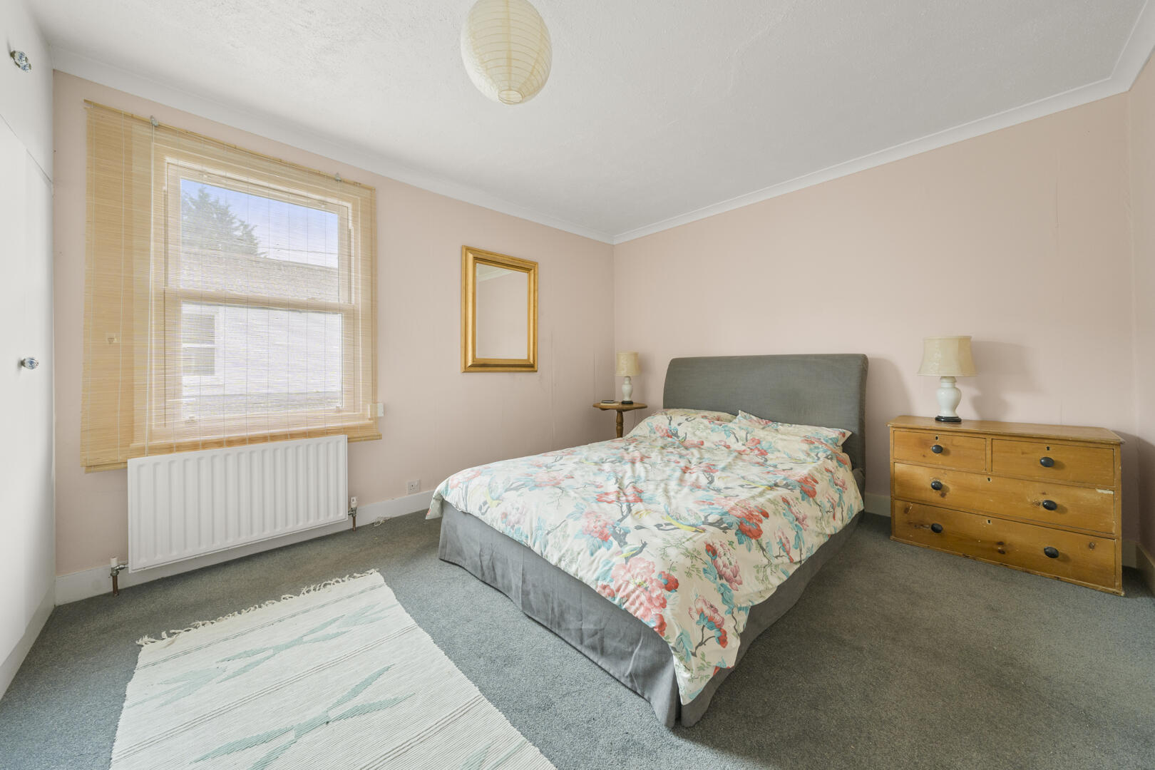 A cozy bedroom with a floral quilted bed, gray upholstered headboard, wooden dresser, and soft pink walls bathed in natural light from a window with blinds.