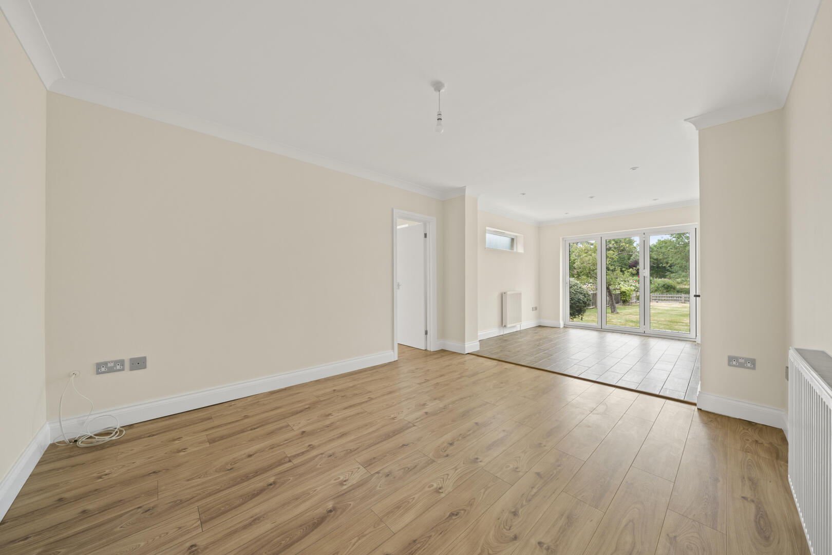Empty living room with light beige walls, white trim, wood-effect flooring, and large glass doors opening to a green garden area.