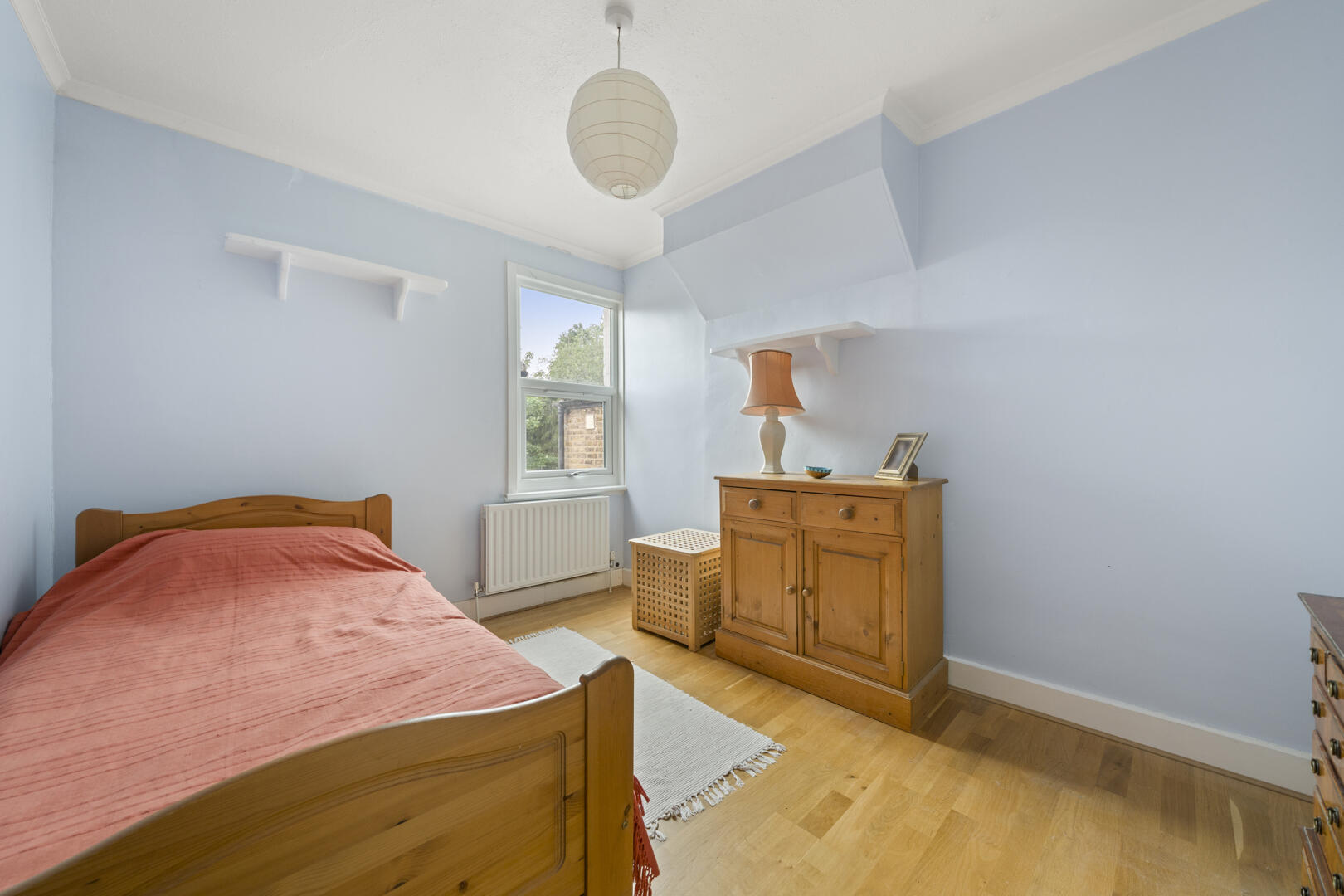Cozy bedroom with pastel blue walls, a single wooden bed with a pink blanket, and a wooden dresser with a lamp and small decor items by the window edge.