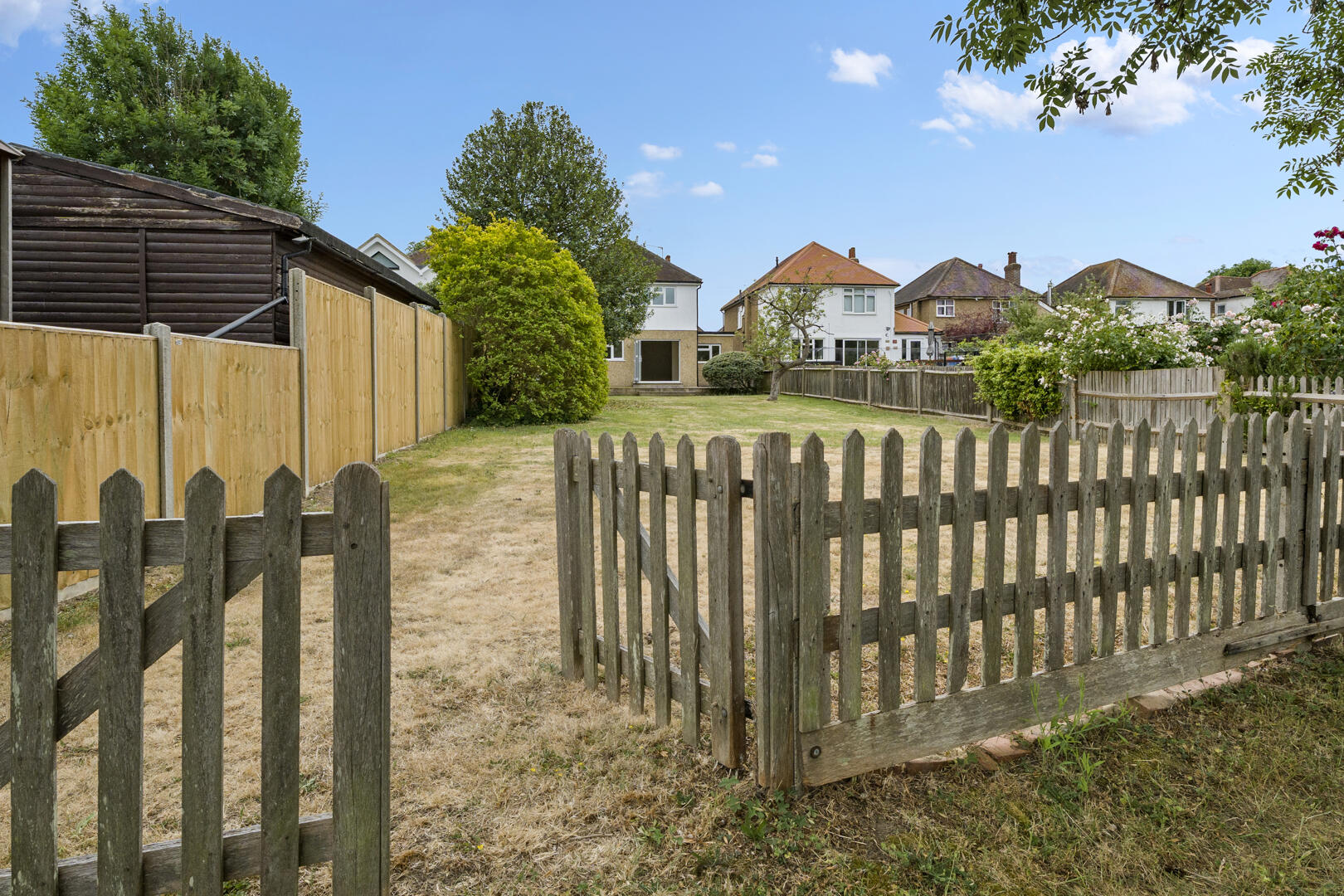Backyard view with a weathered wooden fence and gate, dry grass, and houses in the distance under a blue sky.