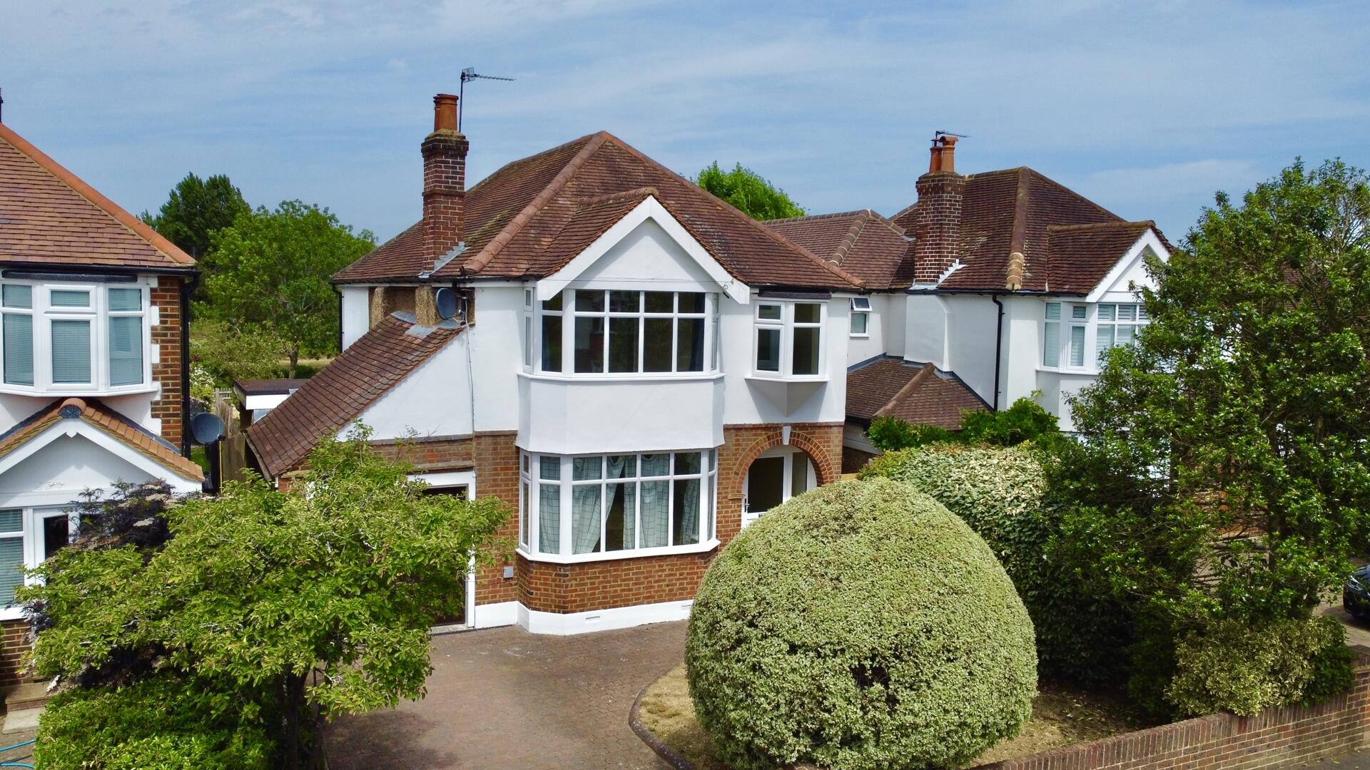 Row of white, red-brick houses with bay windows and red-tiled roofs, flanked by green hedges and trees in a sunny neighborhood court