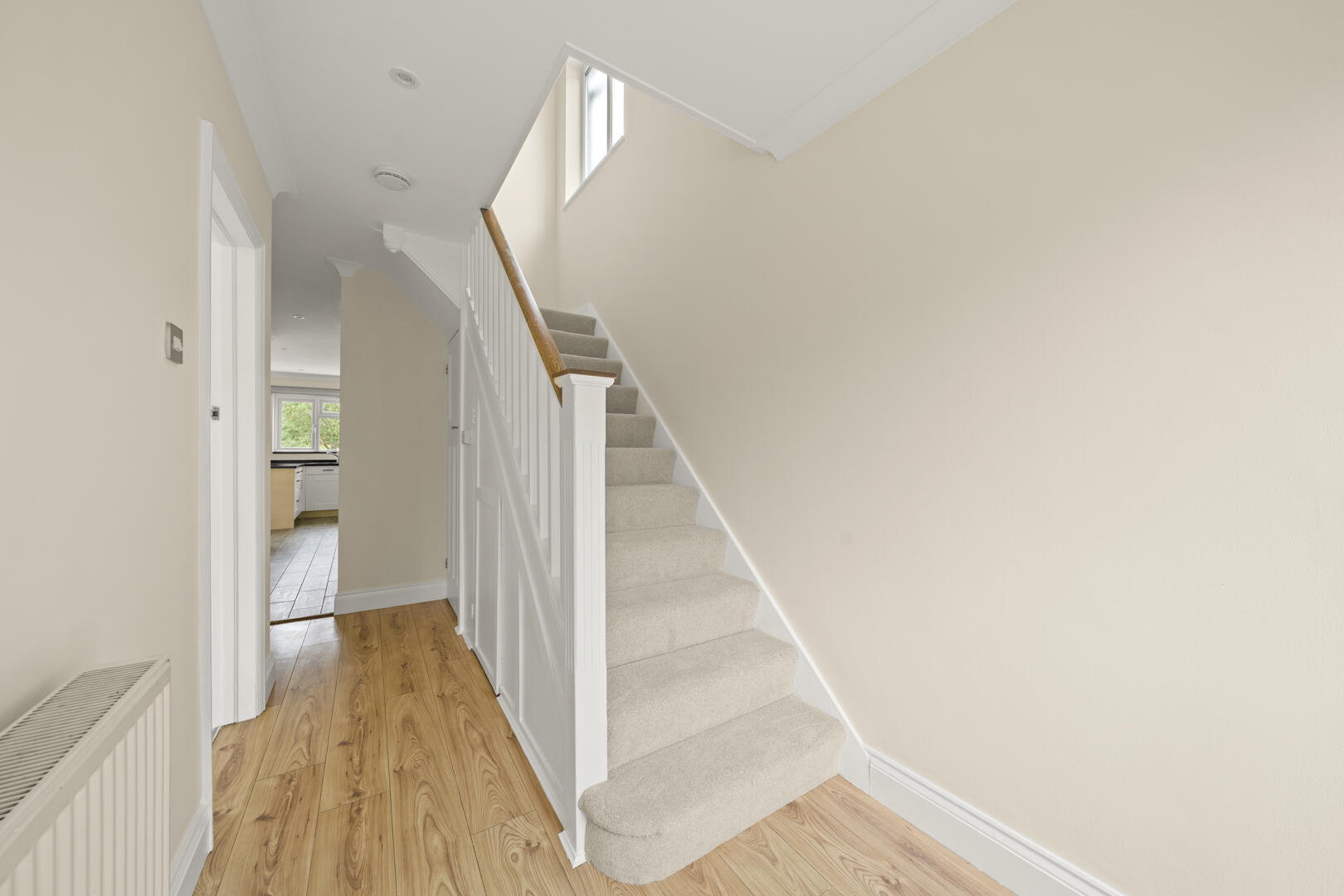 Hallway with beige walls and a carpeted staircase along the right, wooden floor at the bottom, and a kitchen visible in the distance on the left.