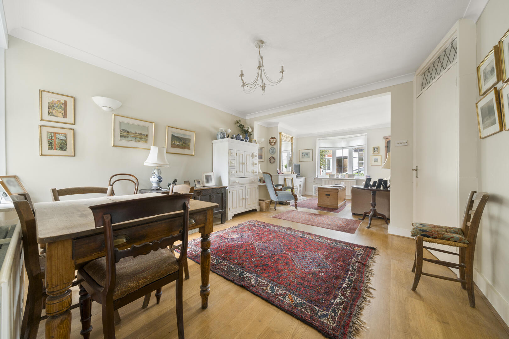 Bright antique living room with wooden dining furniture, oriental rug, and white display cabinet along a pale wall. A chandelier hangs from the ceiling.
