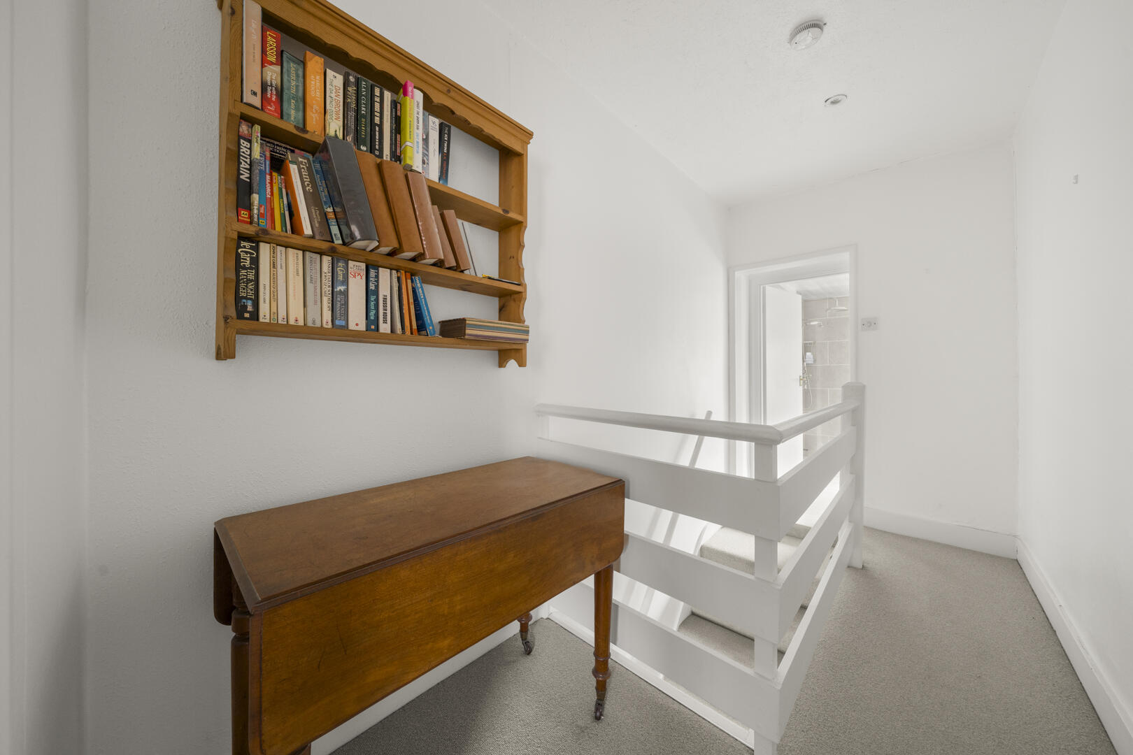 Wooden wall bookcase filled with books above a vintage drop‑leaf table in a bright hallway near a white stair railing.