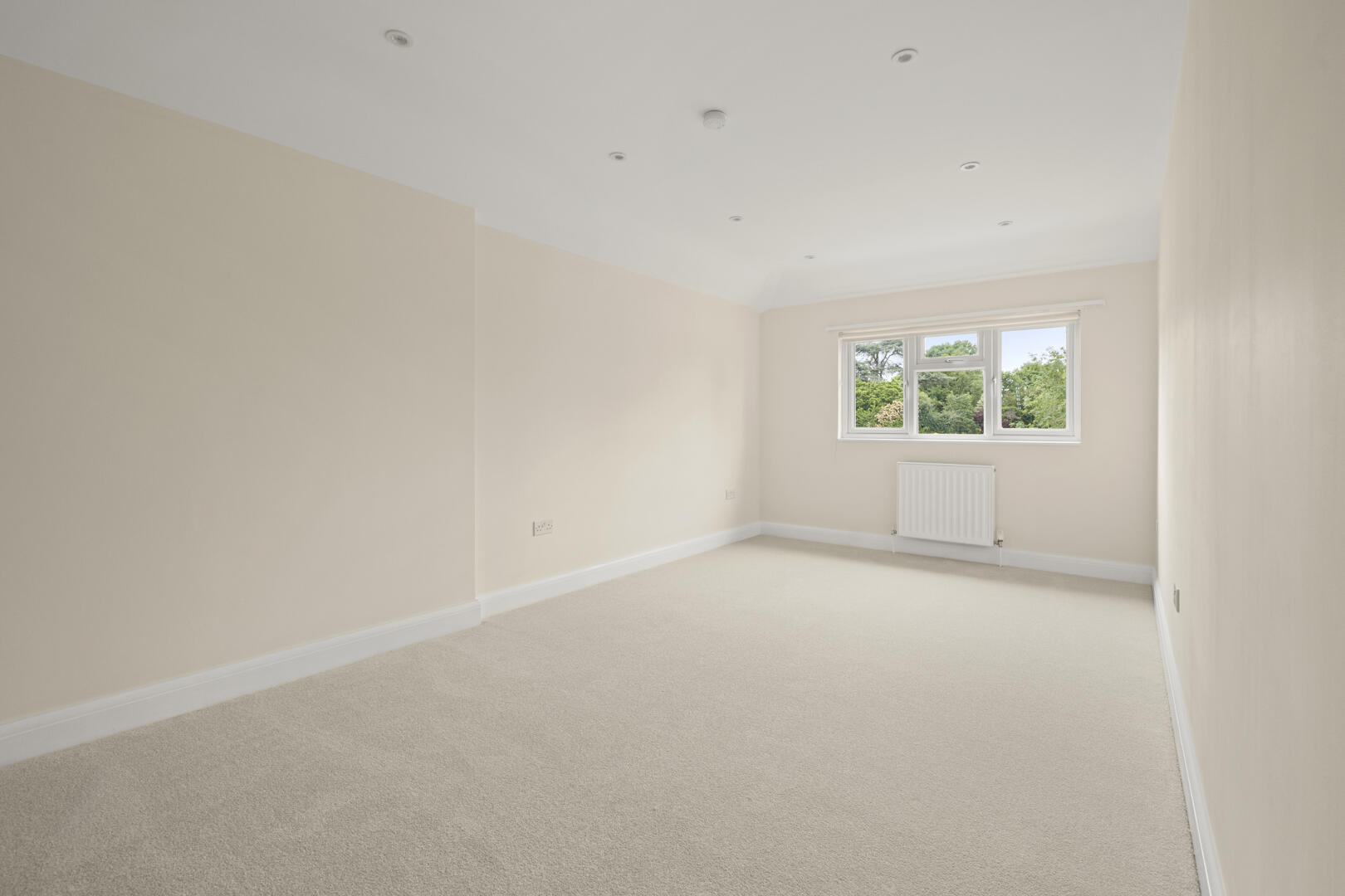 Empty beige, unfurnished room with a large window and radiator on the wall; cream carpet and recessed ceiling lights','Unoccupied light-colored room with cream walls, a white radiator under a window, and a beige carpet','Bright, empty room featuring a bay of windows, radiator, and subtle ceiling spotlights on a pale backdrop','Neutral-toned, empty living space with a window view, white baseboards, and a soft carpet finish','Spacious, minimal room with beige walls, white trim, and a large window overlooking greenery