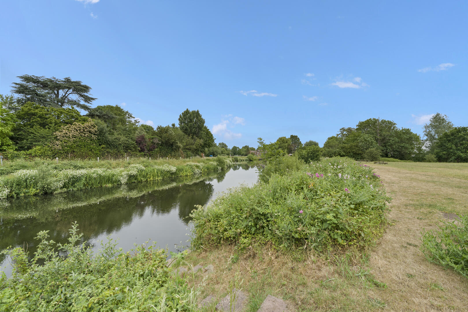 Calm canal lined with green shrubs and trees under a clear blue sky in a park setting.