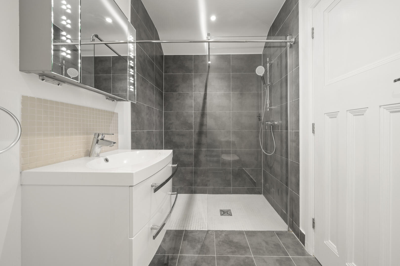 Modern bathroom with a white vanity and curved sink, beige tiled backsplash, and mirrored medicine cabinet above the sink.