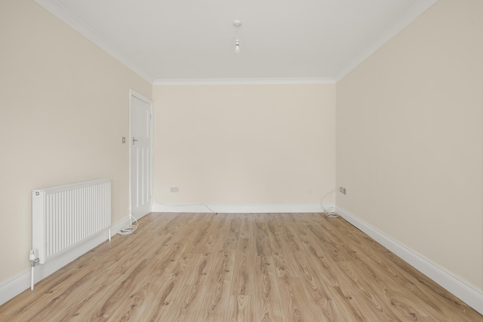 Empty beige room with a light wood floor, radiator on the left, white door, and exposed cables along the baseboard.