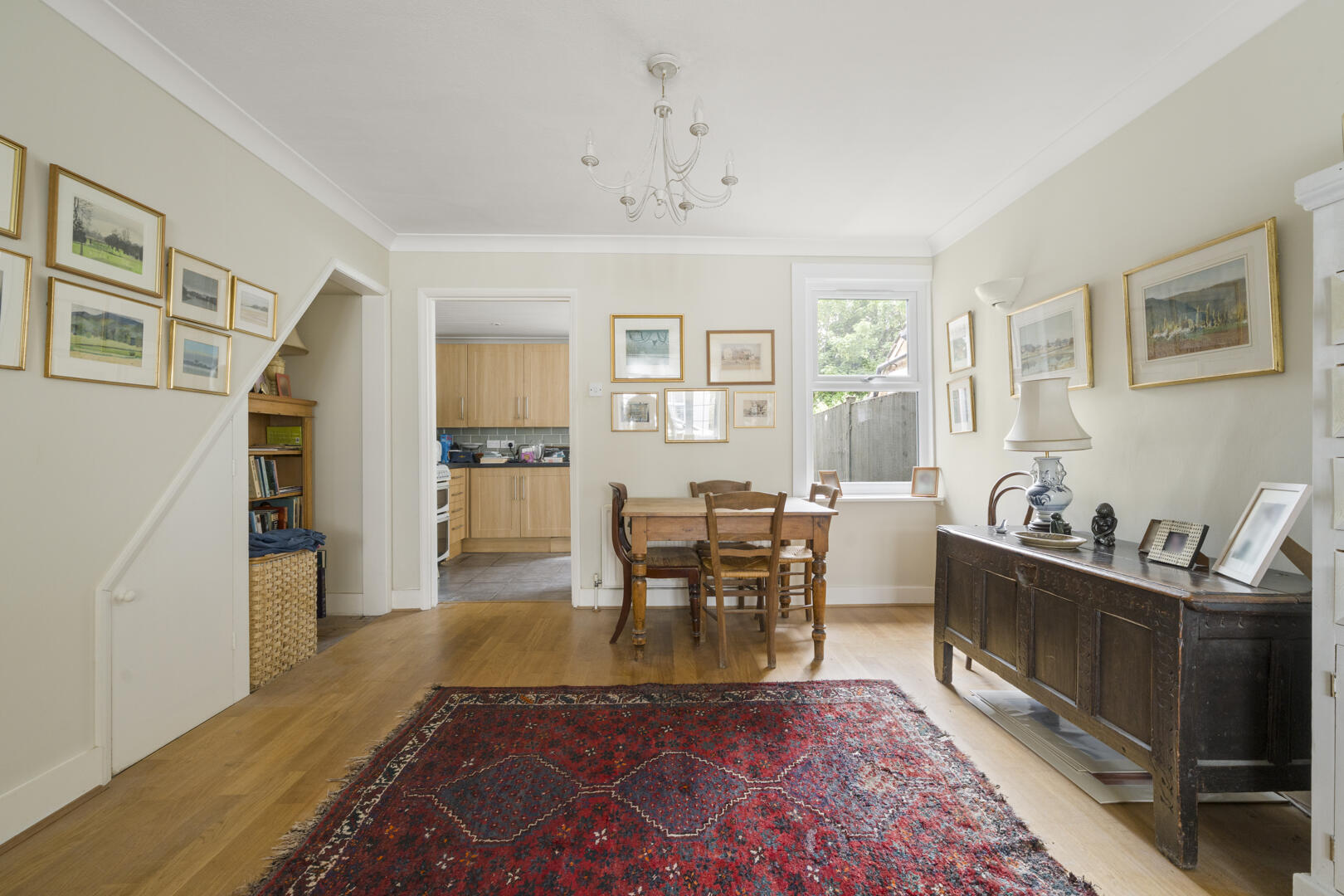 Dining room with a wooden table and chairs on a red patterned rug, pale walls, and many framed pictures above the sideboard and window.