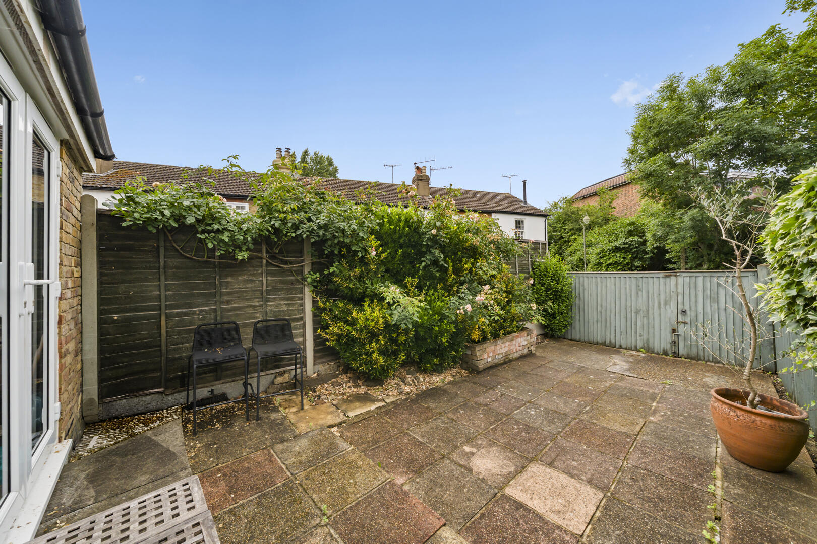 Small backyard patio with tiled concrete slabs, a wooden fence, climbing greenery, and two black metal chairs along the fence; a large potted tree on the right and a blue sky above.