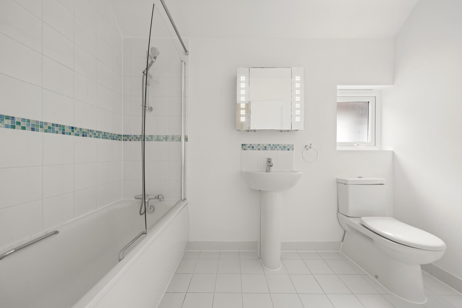 White minimalist bathroom with a tub and glass shower screen, mosaic tile accent, pedestal sink, lighted mirror, and toilet near a window