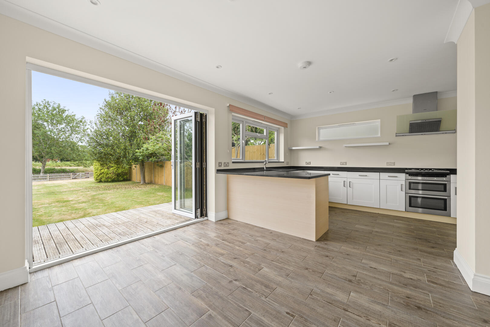 Open-plan kitchen with an island, white cabinets, and black countertops, stainless steel oven and range hood, overlooking a backyard through large glass doors.