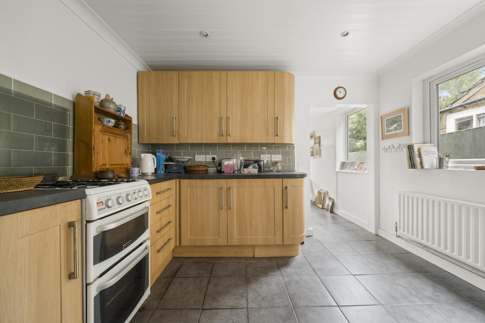 Bright kitchen with light wood cabinets, grey tile backsplash, and a white freestanding oven along the left wall; window and radiator visible on the right.