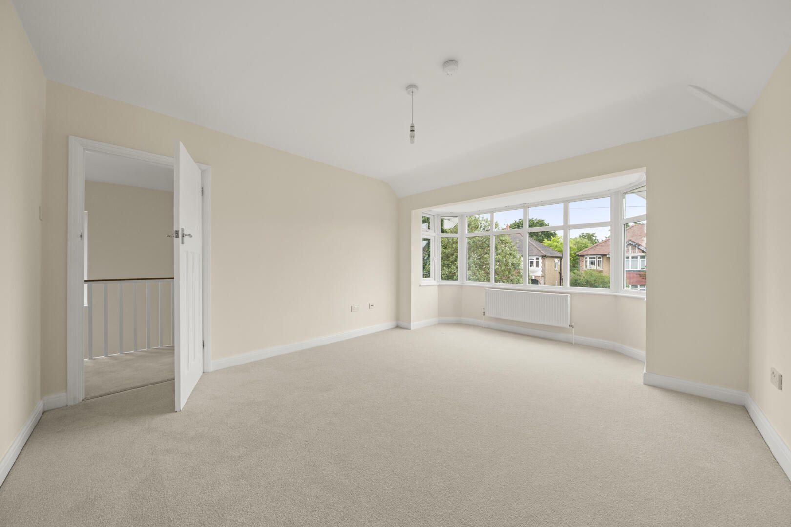 Bright empty living room with a bay window, cream walls, radiator, and an open door to the stairs area.