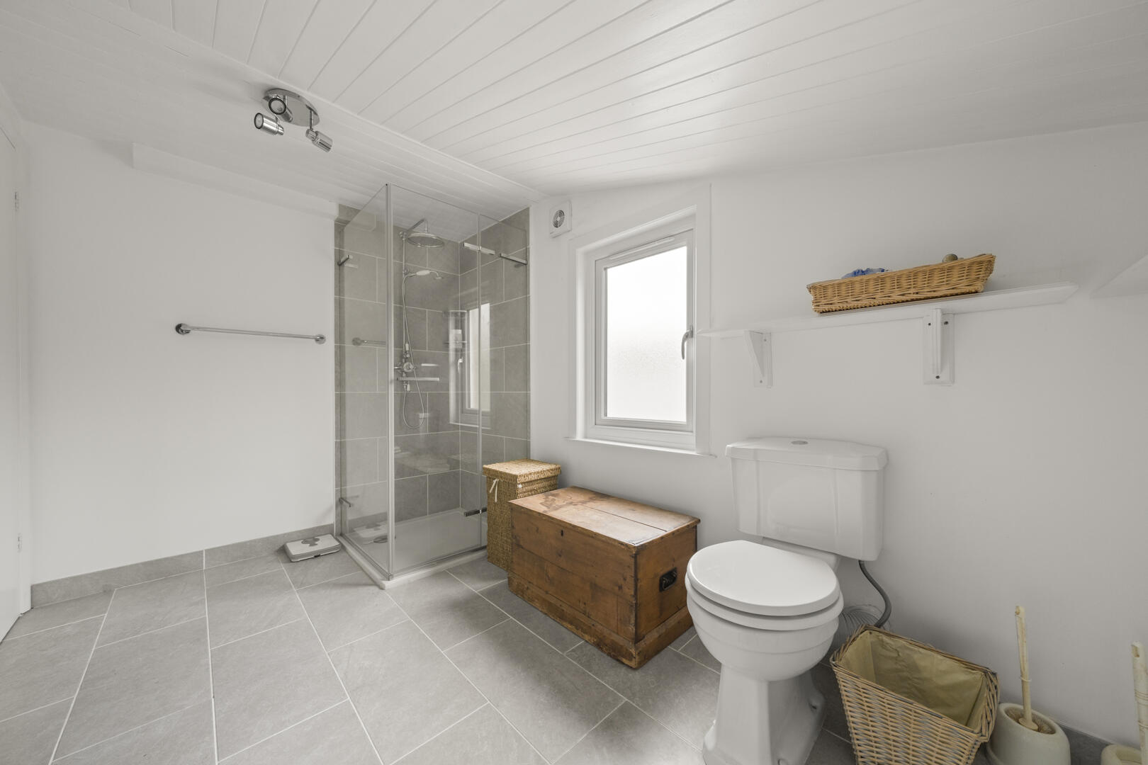 Bright bathroom with a glass-walled shower, toilet, and a wooden chest beside the window on gray tiled floor