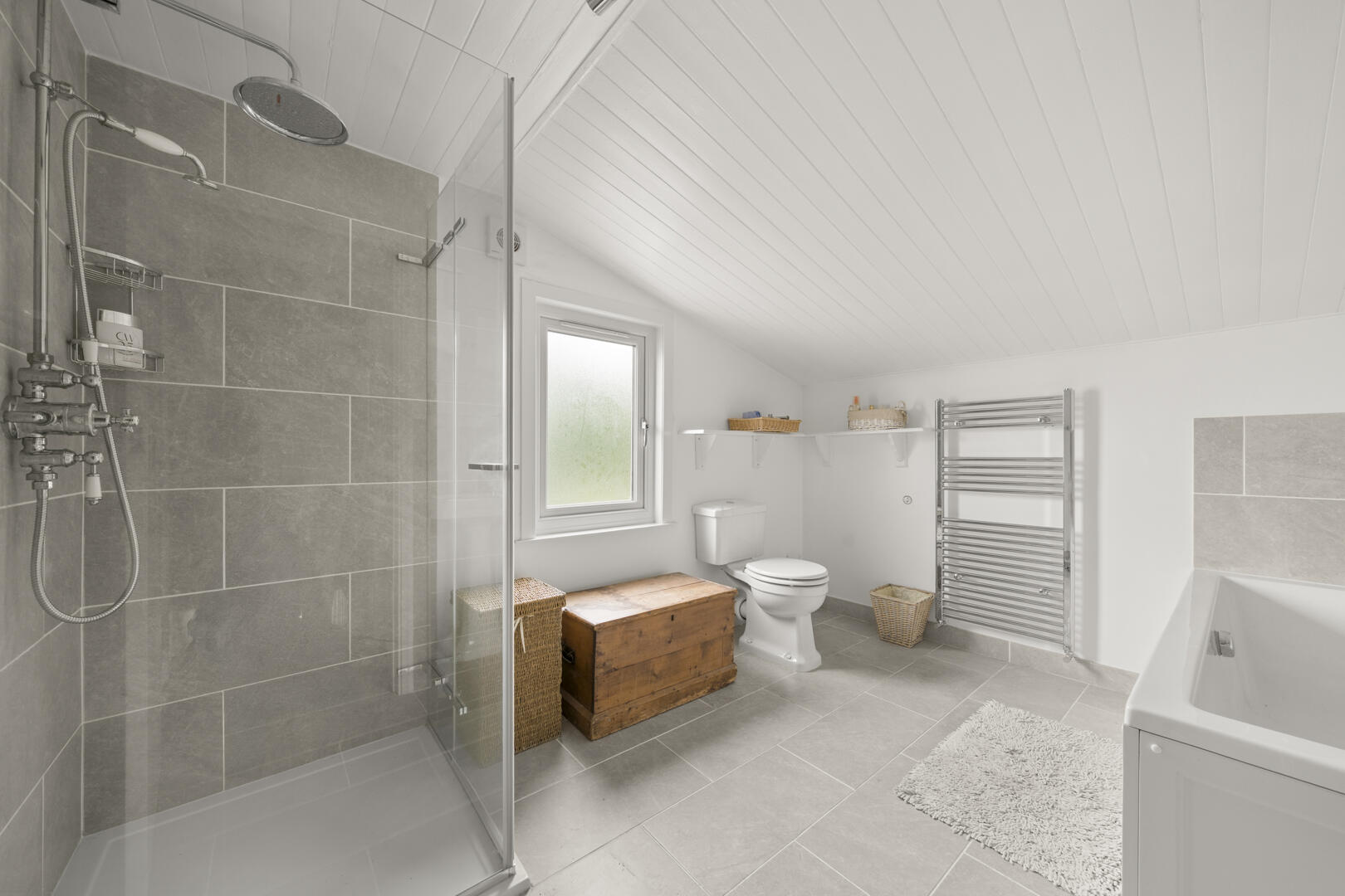 Bright modern bathroom with a glass-enclosed shower, gray tiled walls, white toilet and window, plus a wooden chest and wicker baskets on the floor area.