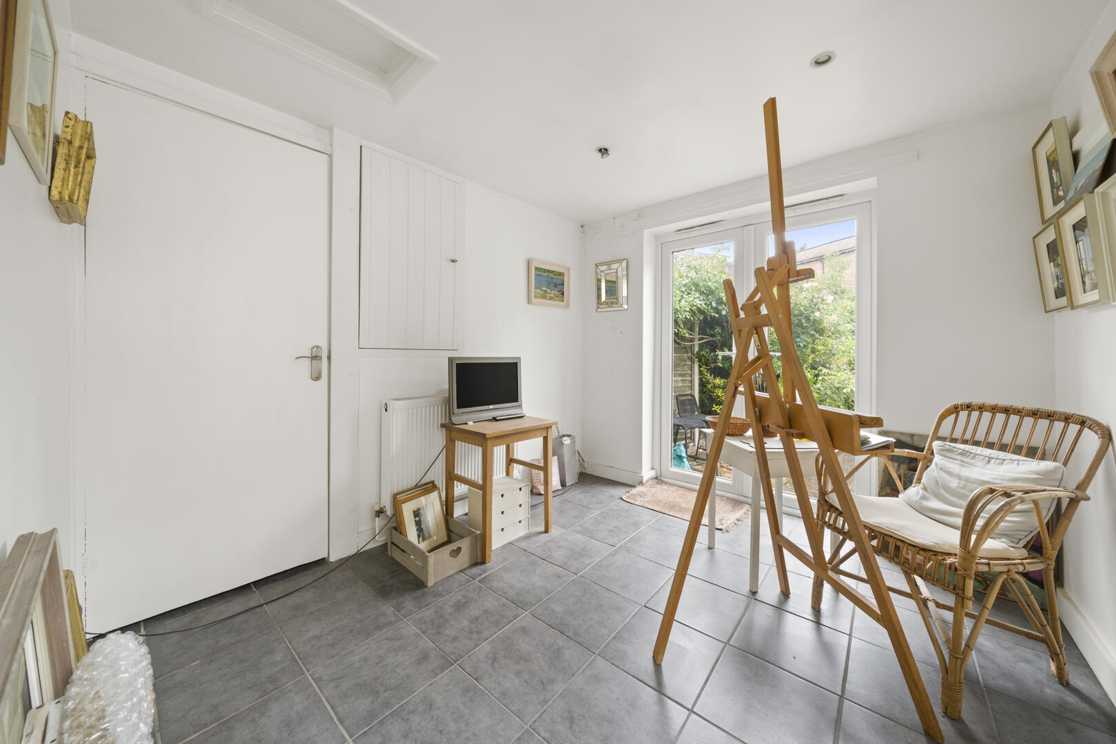 Bright white room with a wooden easel near large glass doors to a garden and a wicker chair nearby you can see framed art on the wall.