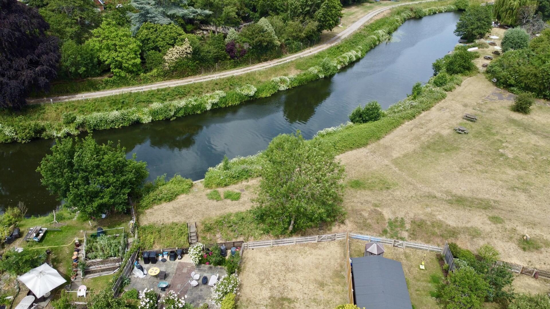 Aerial view of a winding river bordered by trees, with a dirt path along the bank and a grassy park area with picnic tables.