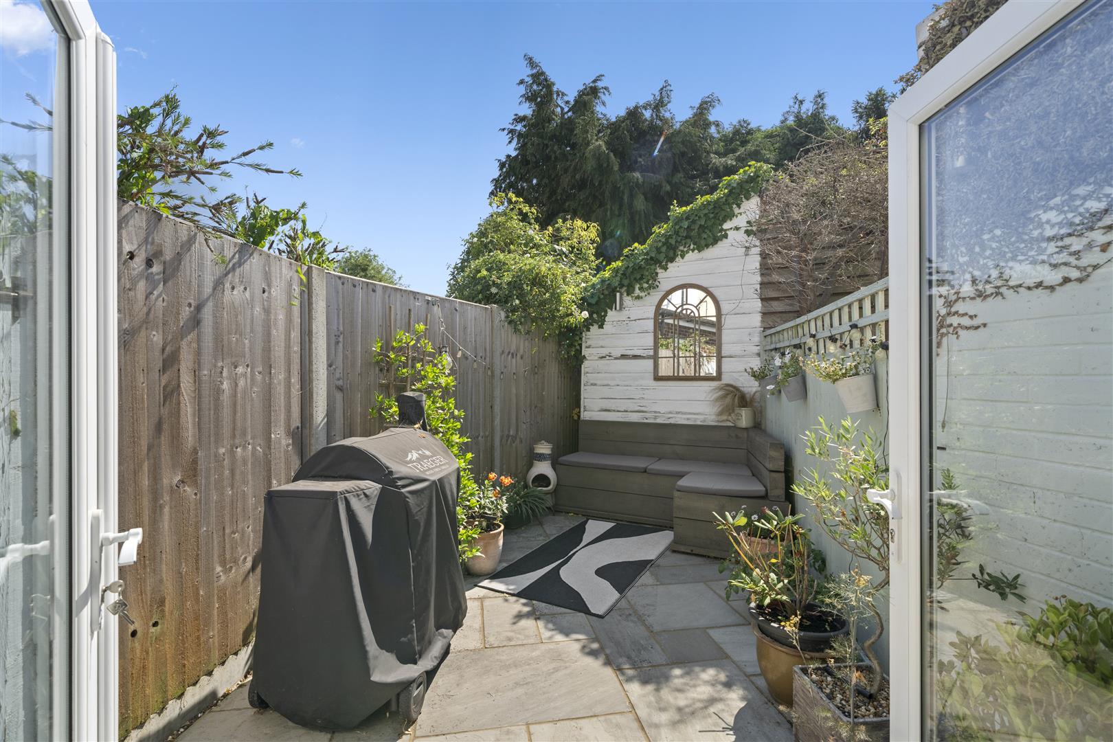Small outdoor patio with a covered grill, gray sectional seating, potted plants, and ivy-covered walls beside a white shed.