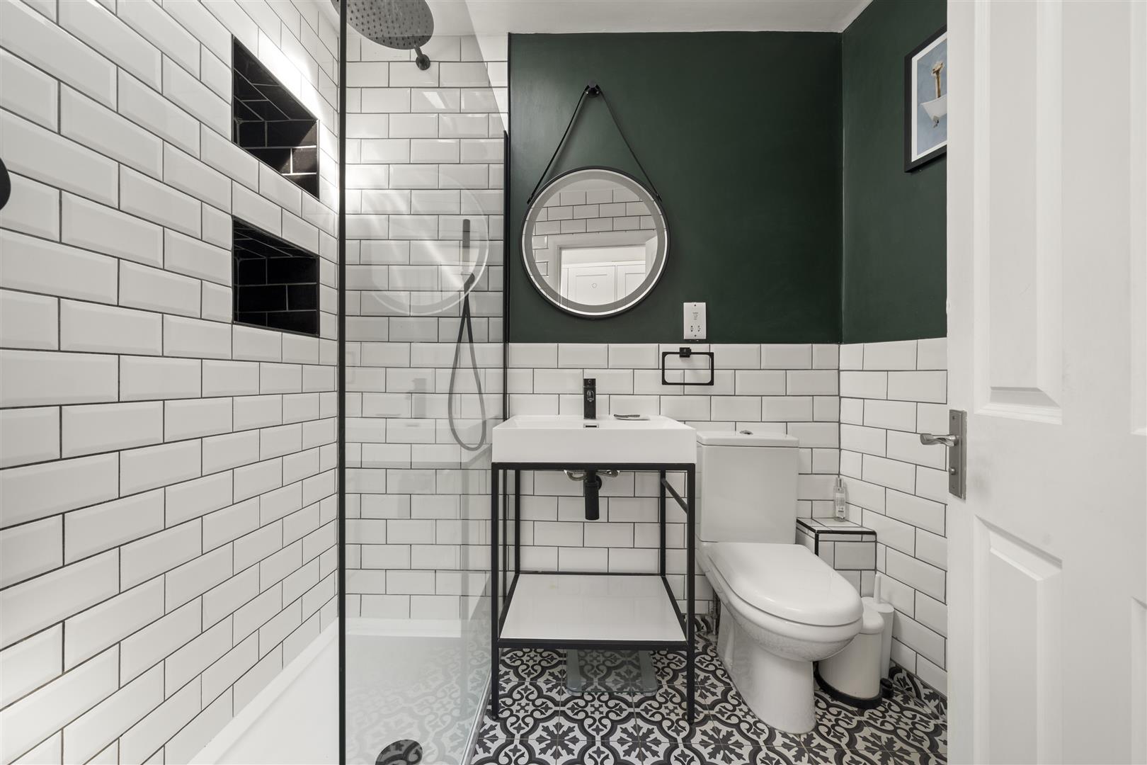 Modern bathroom with white subway tiles, dark green accent wall, round mirror, and a white sink on a black metal stand with a patterned floor suburban style.