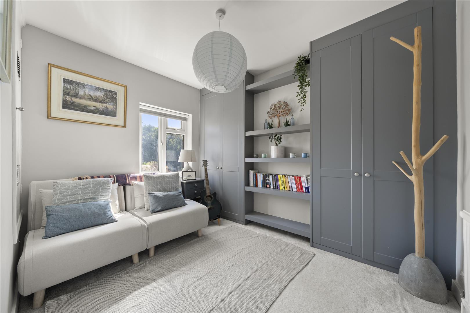 Cozy living room with gray built-in wardrobes, open shelves, and a beige sofa with blue cushions by a window and guitar beside it.